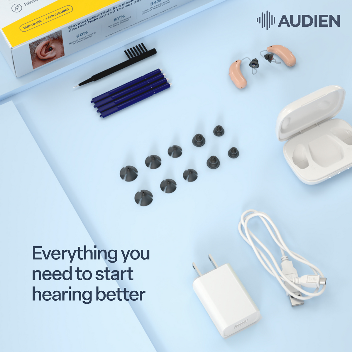 Audien hearing assistance kit laid out on a light blue background, including beige in-ear hearing devices, a white charging case, a white wall charger, a USB cable, black replacement ear tips, four blue cleaning tools, a small brush, and the product box. The Audien logo appears in the top right, and text reads 'Everything you need to start hearing better'.